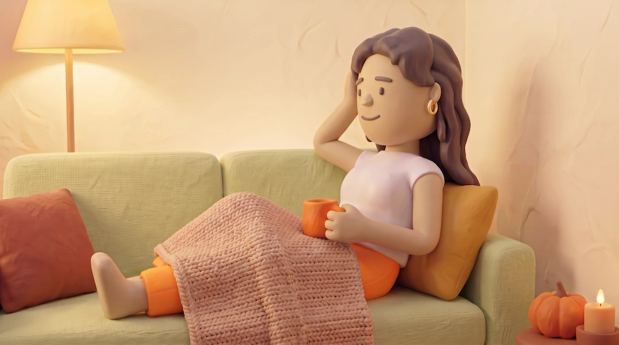 Person sitter bekvämt i en soffa med en filt över benen och en kopp i handen. En golvlampa lyser upp rummet och höstdekorationer med pumpa och ljus syns i bakgrunden.